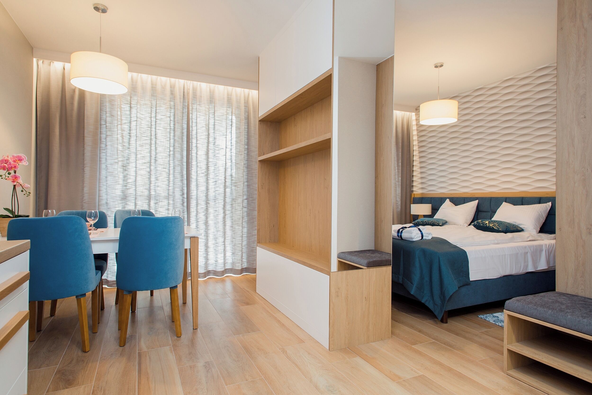 A modern hotel room with a wooden floor, a bed with a blue bedspread, a desk with chairs, and a dining area with a table and chairs.
