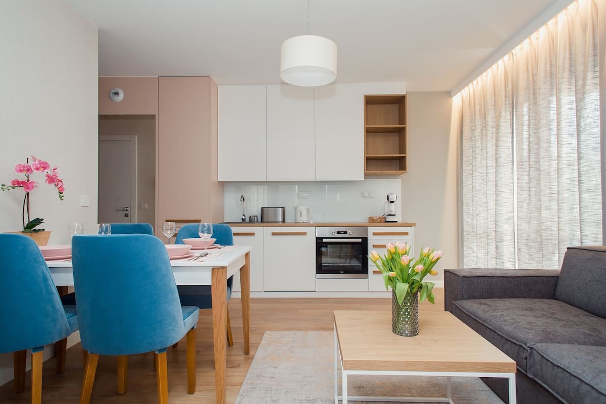 A modern living room with a sofa, a dining table with chairs, a kitchen area with cabinets, and a window with curtains.