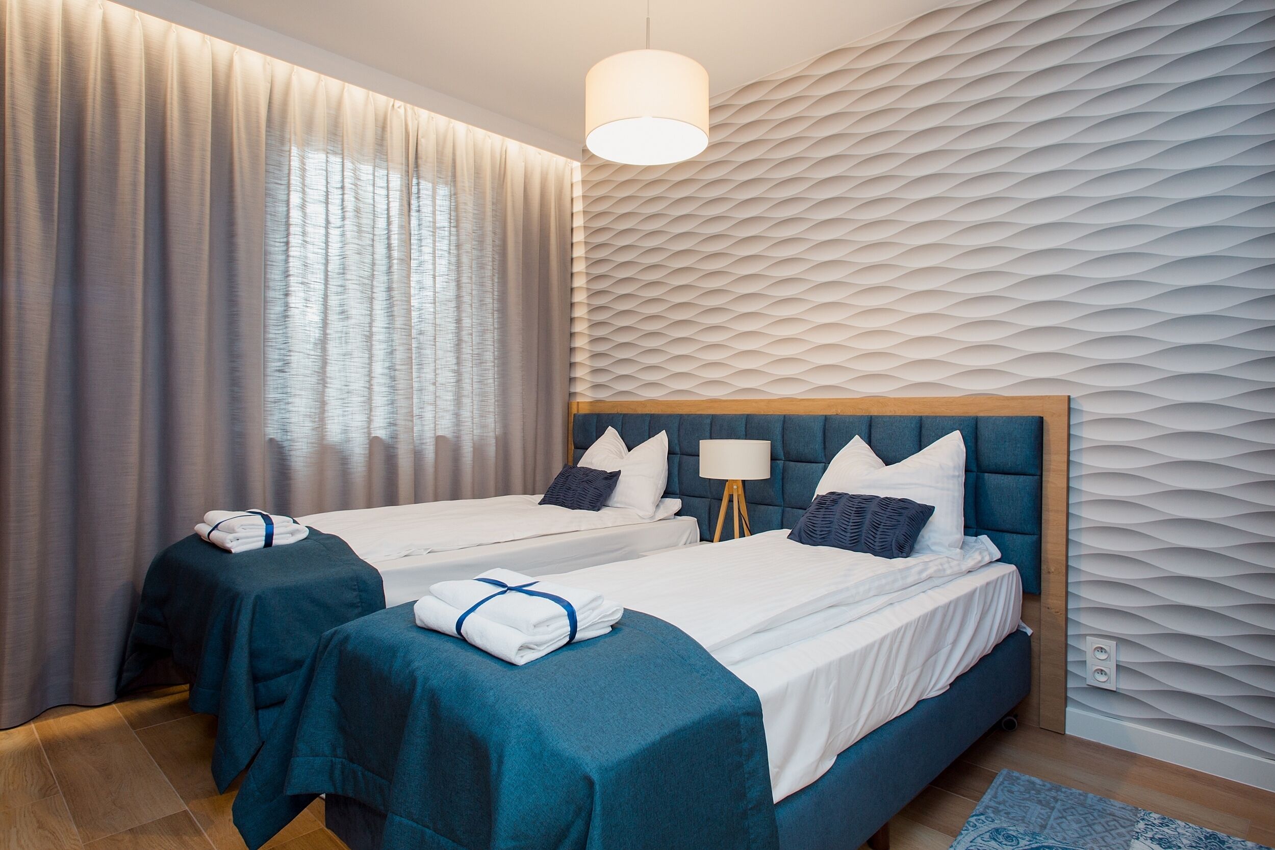 A hotel room with two beds, a large window with curtains, a textured wall, and a hanging light fixture.