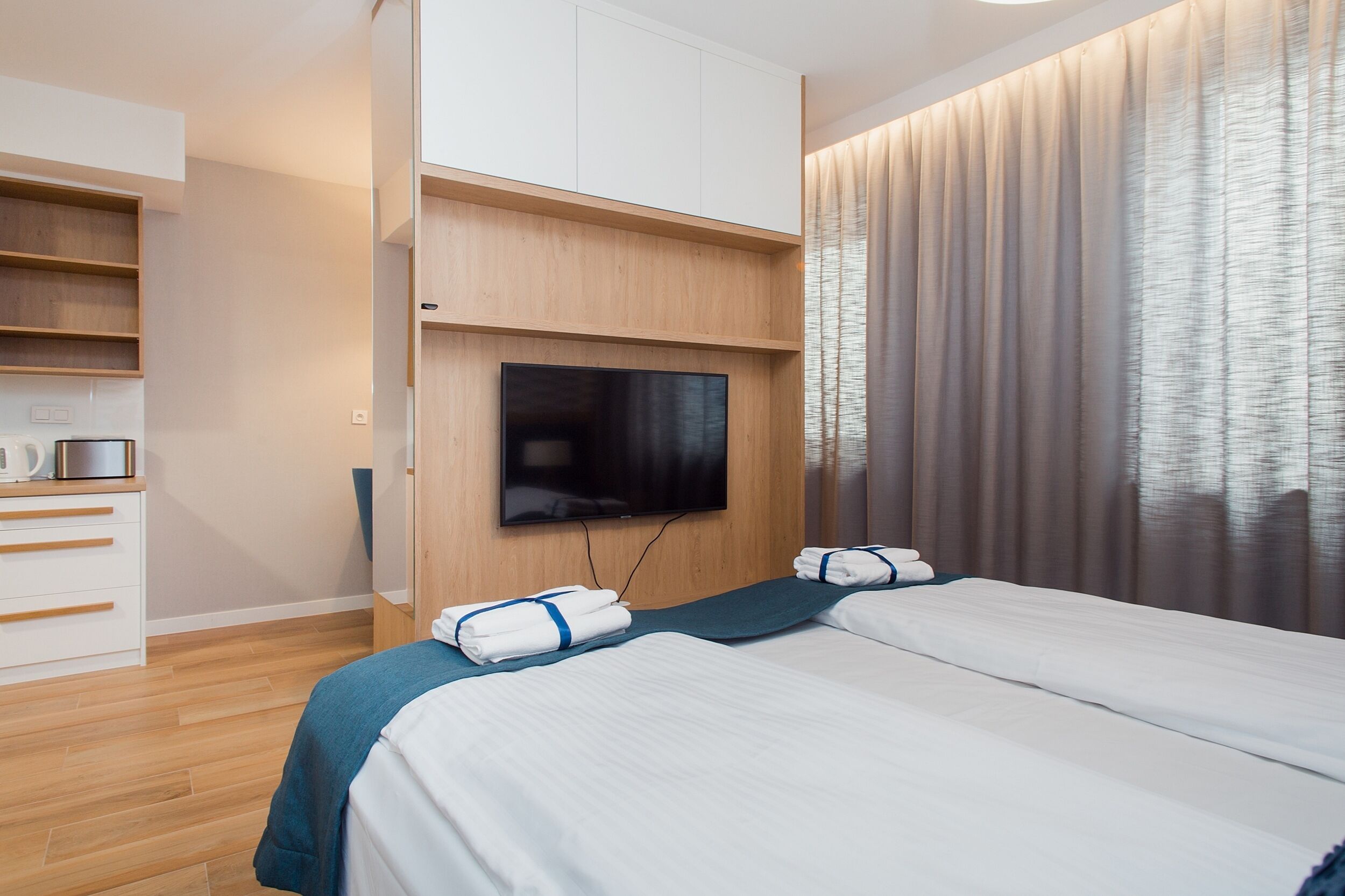 Habitación de hotel con cama, televisor montado en la pared, armario de madera y ventana con cortinas.