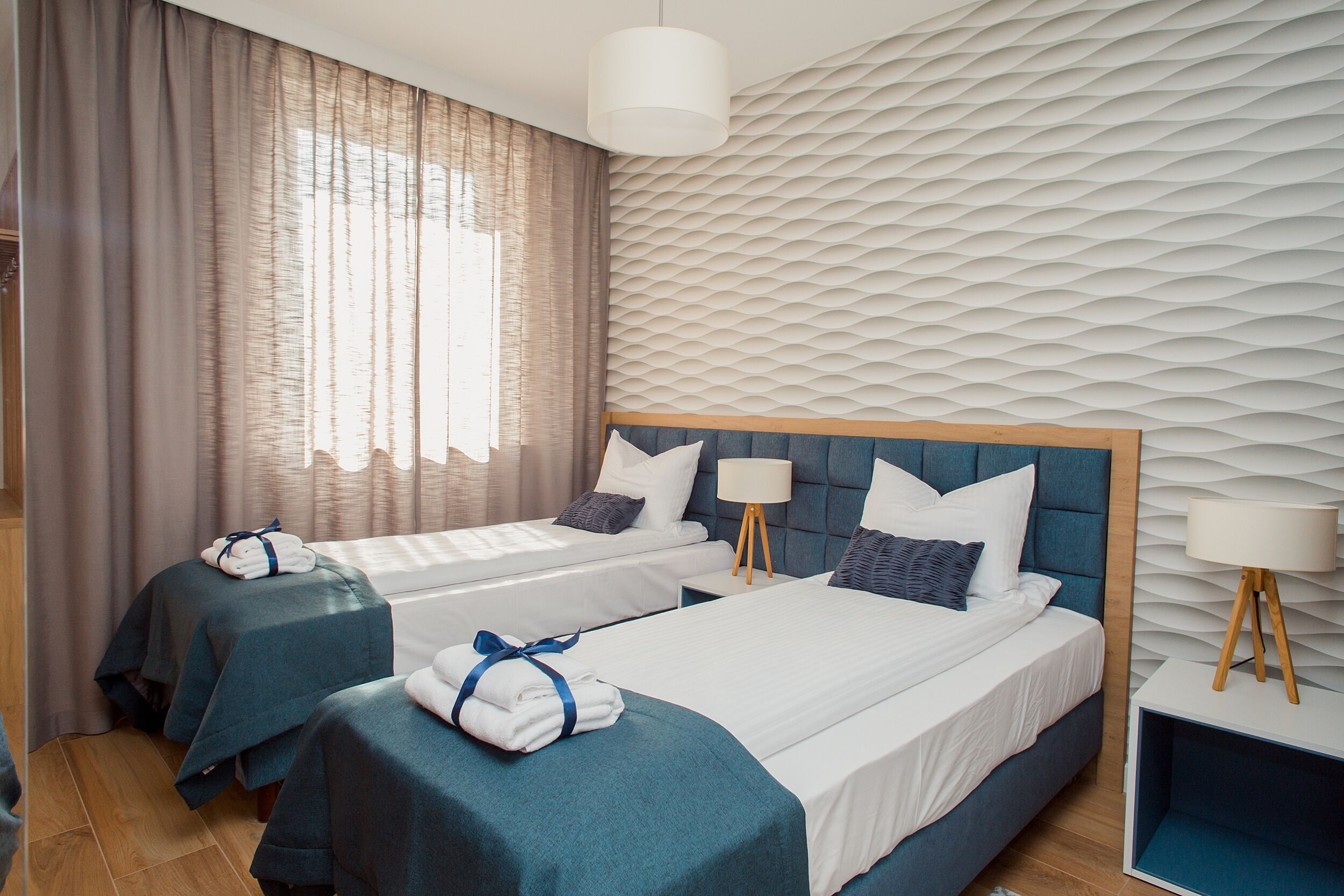 A hotel room with two beds, a large window with curtains, a textured wall, and a bedside table with a lamp.