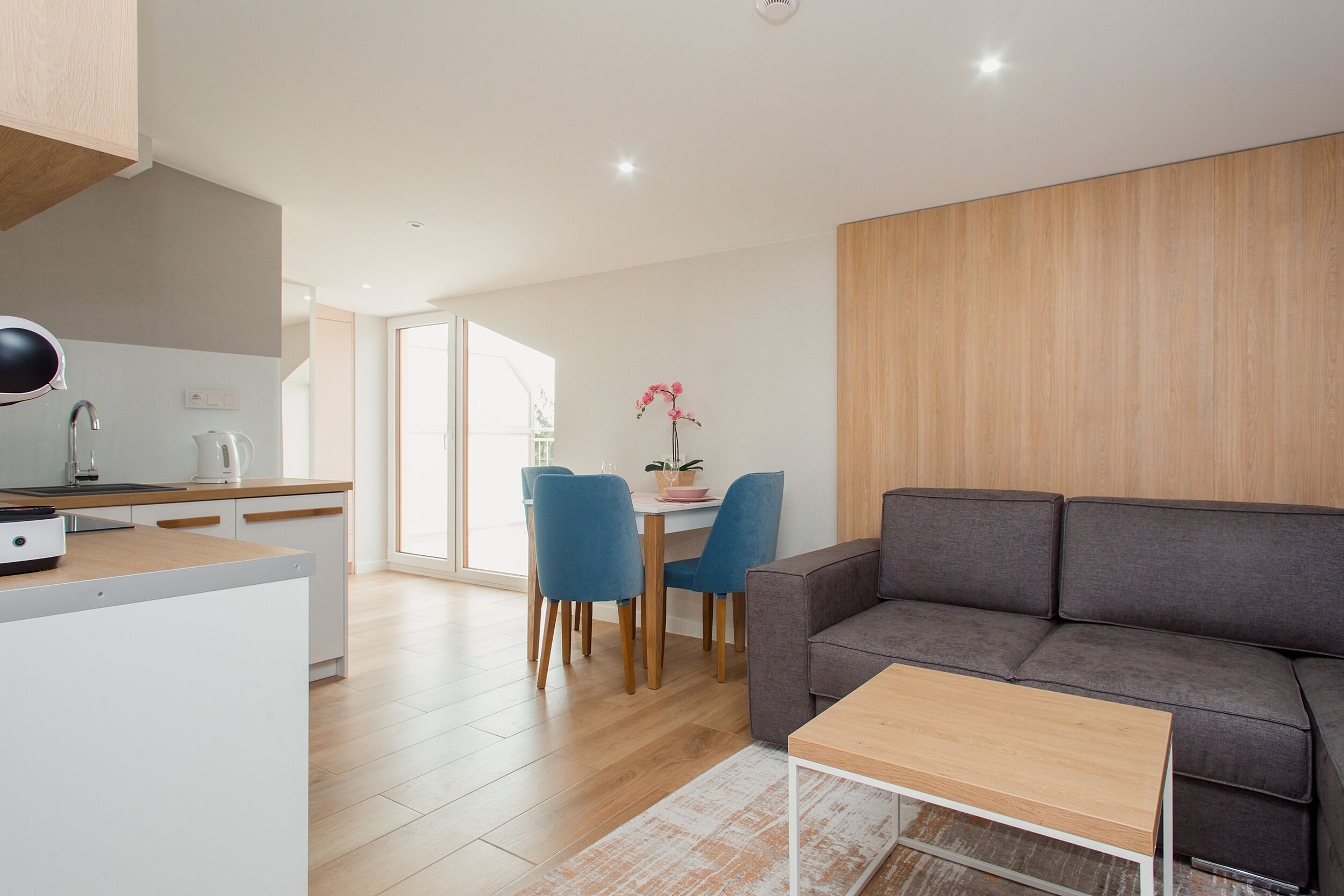 A modern living room with a grey sofa, wooden coffee table, and a kitchen area with a white countertop and blue chairs.