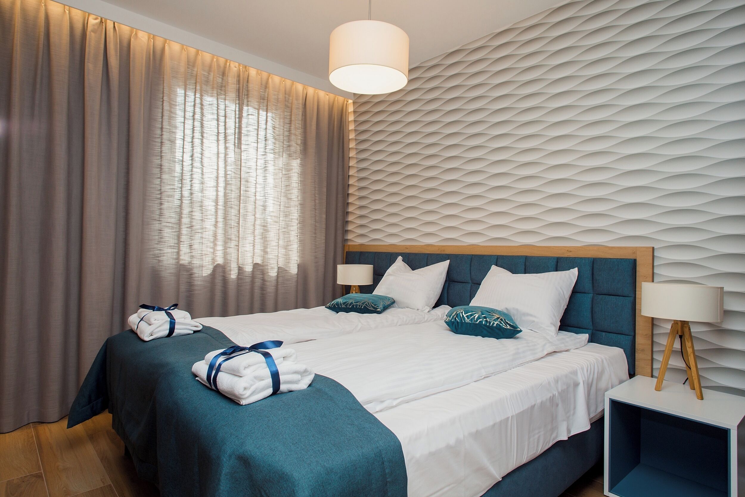 Ein Hotelzimmer mit einem großen Bett, zwei Nachttischen, einer Wand mit Strukturmuster und einem großen Fenster mit Vorhängen.