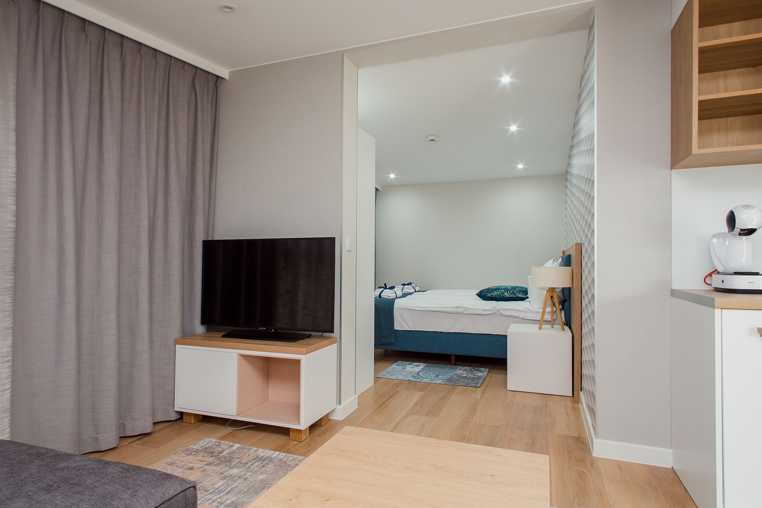 Una camera d'albergo moderna con TV a schermo piatto, un letto con poggiatesta blu, un comodino con una lampada e un angolo cottura.