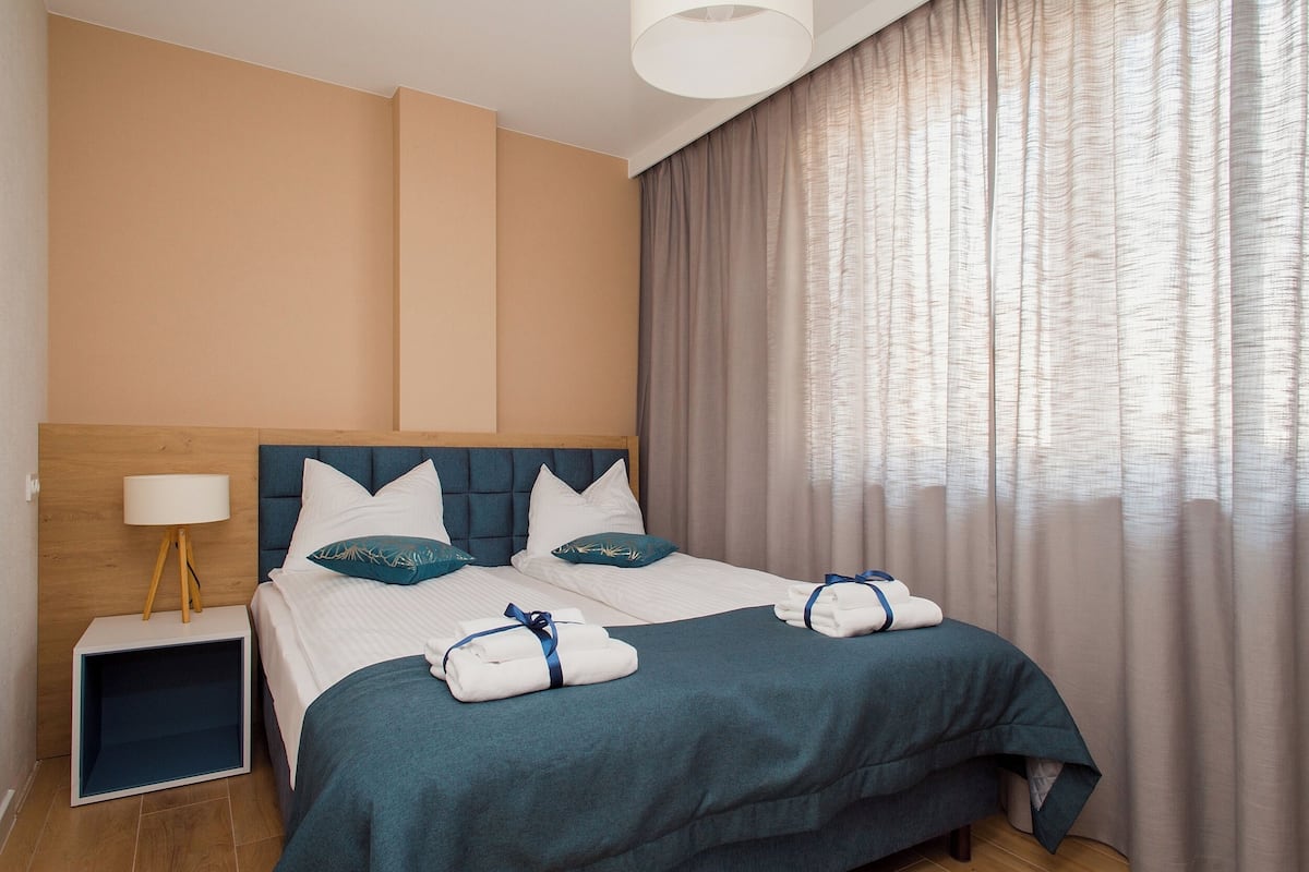 A neatly made bed with a blue headboard, white pillows, and a teal throw blanket. There is a small bedside table with a lamp and a folded towel. The room has beige walls and a large window with sheer curtains.