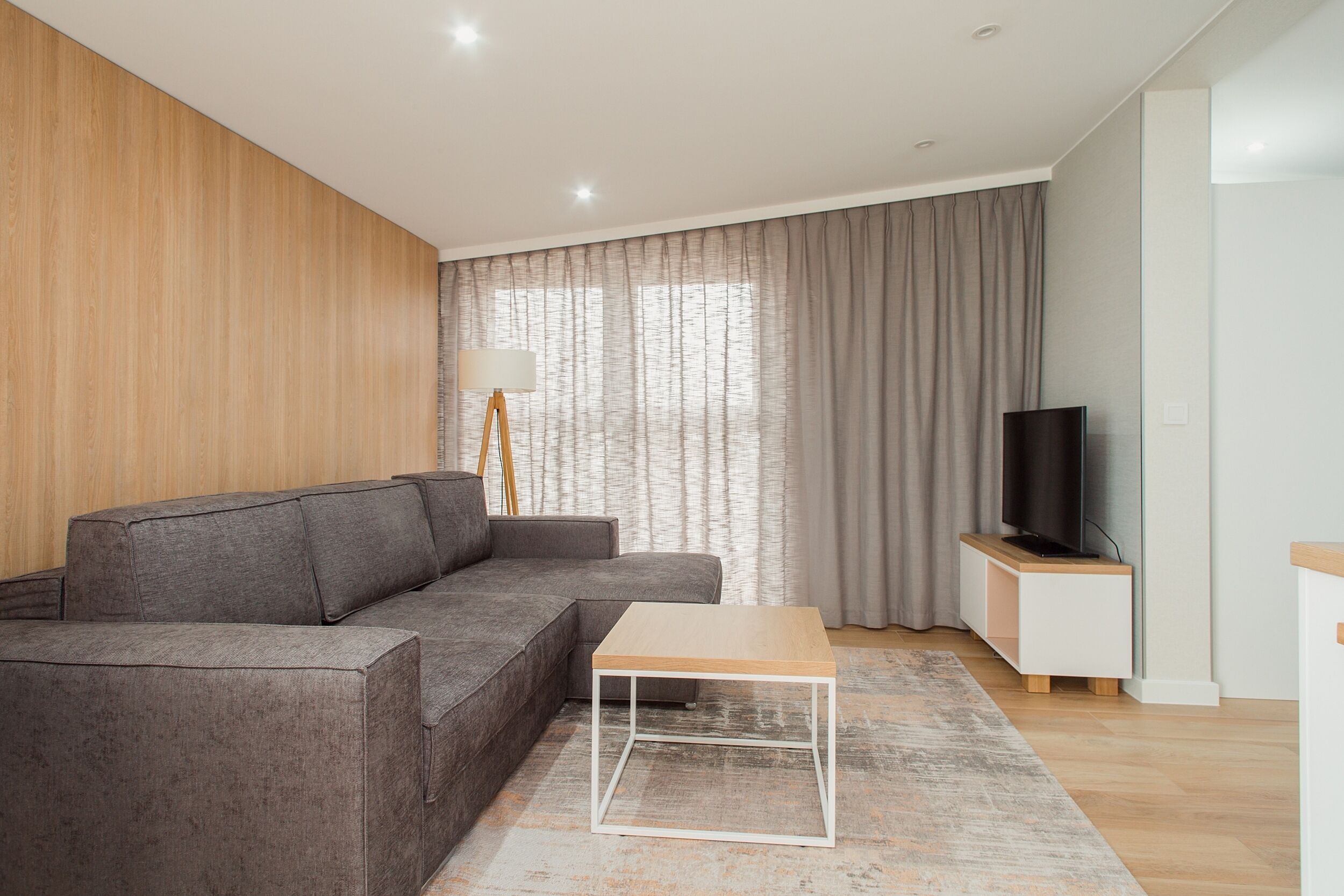 A modern living room with a grey sectional sofa, a wooden coffee table, a floor lamp, a TV on a stand, and a large window with curtains.