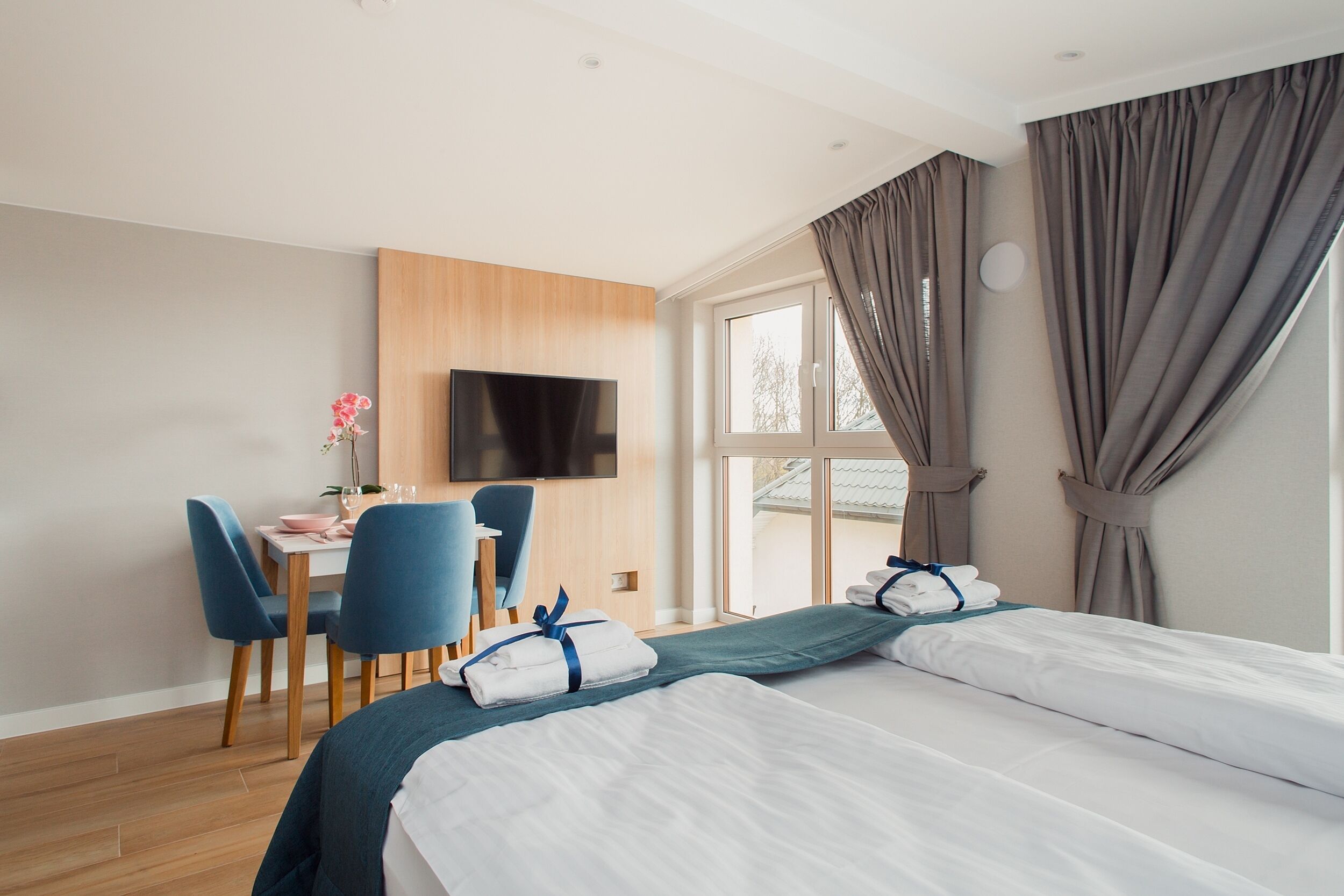 A hotel room with two beds, a desk with chairs, a TV, and large windows with curtains.