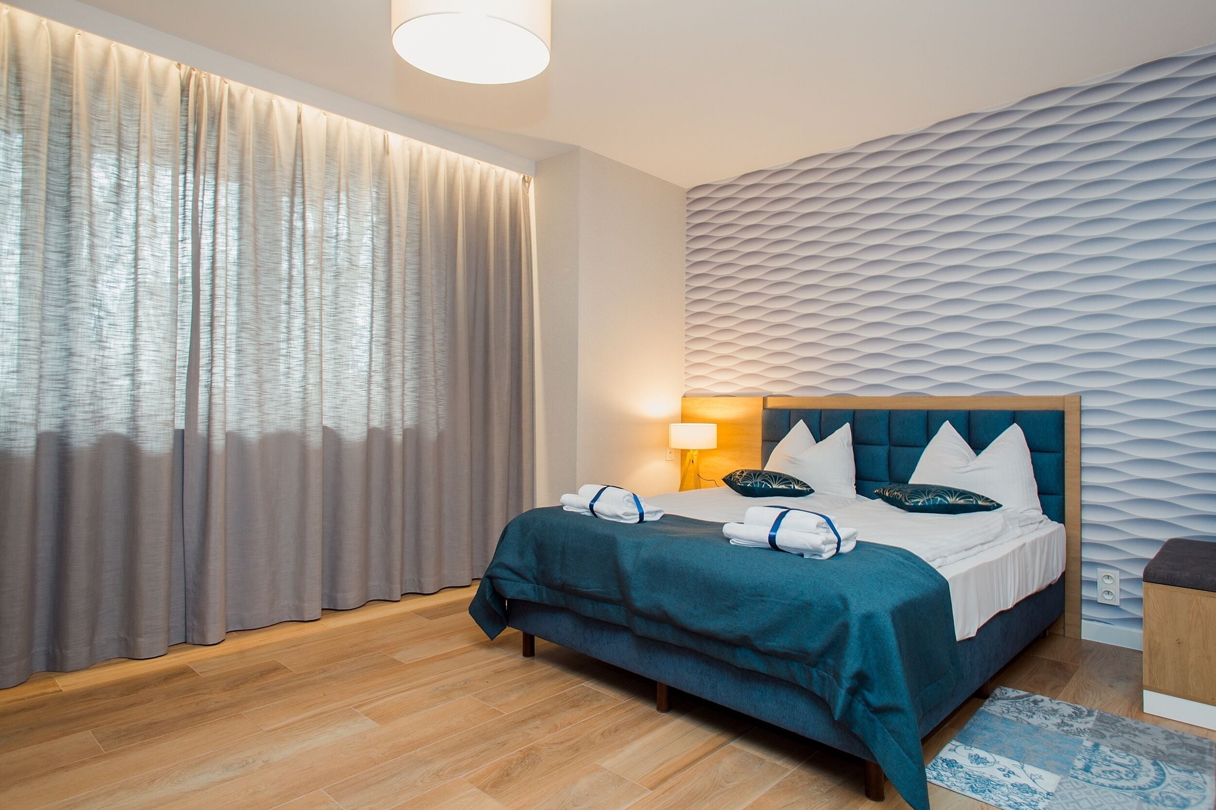 A modern hotel room with a large bed, a headboard with a textured pattern, a bedside lamp, and a wooden floor.
