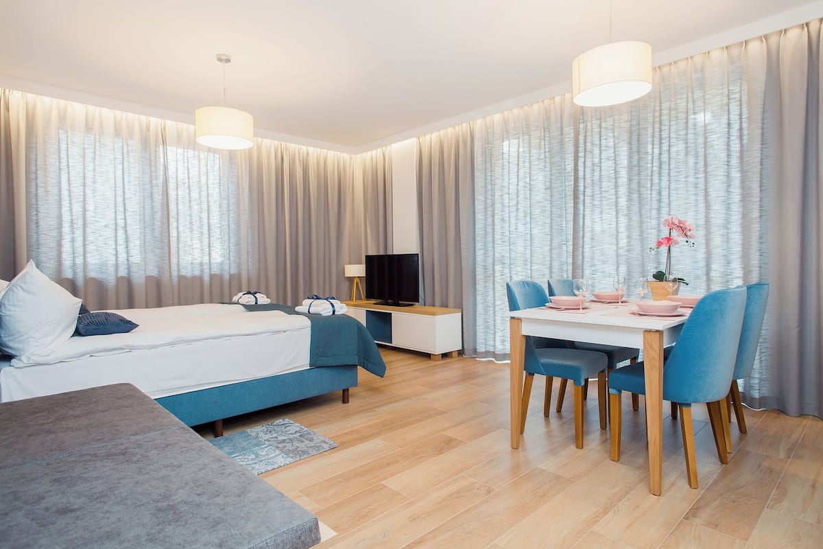 A modern hotel room with a bed, bedside table, TV, and a dining area with chairs and a table.