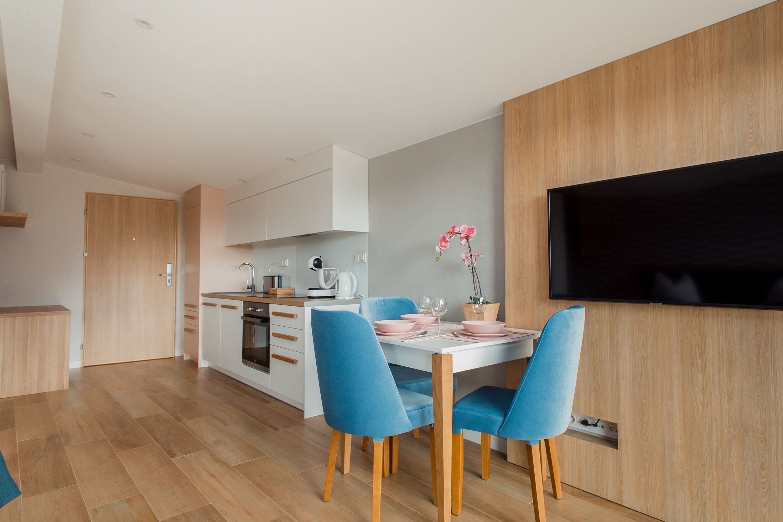 Een moderne keuken met witte kasten, een inbouwoven en een eethoek met een witte tafel en blauwe stoelen.