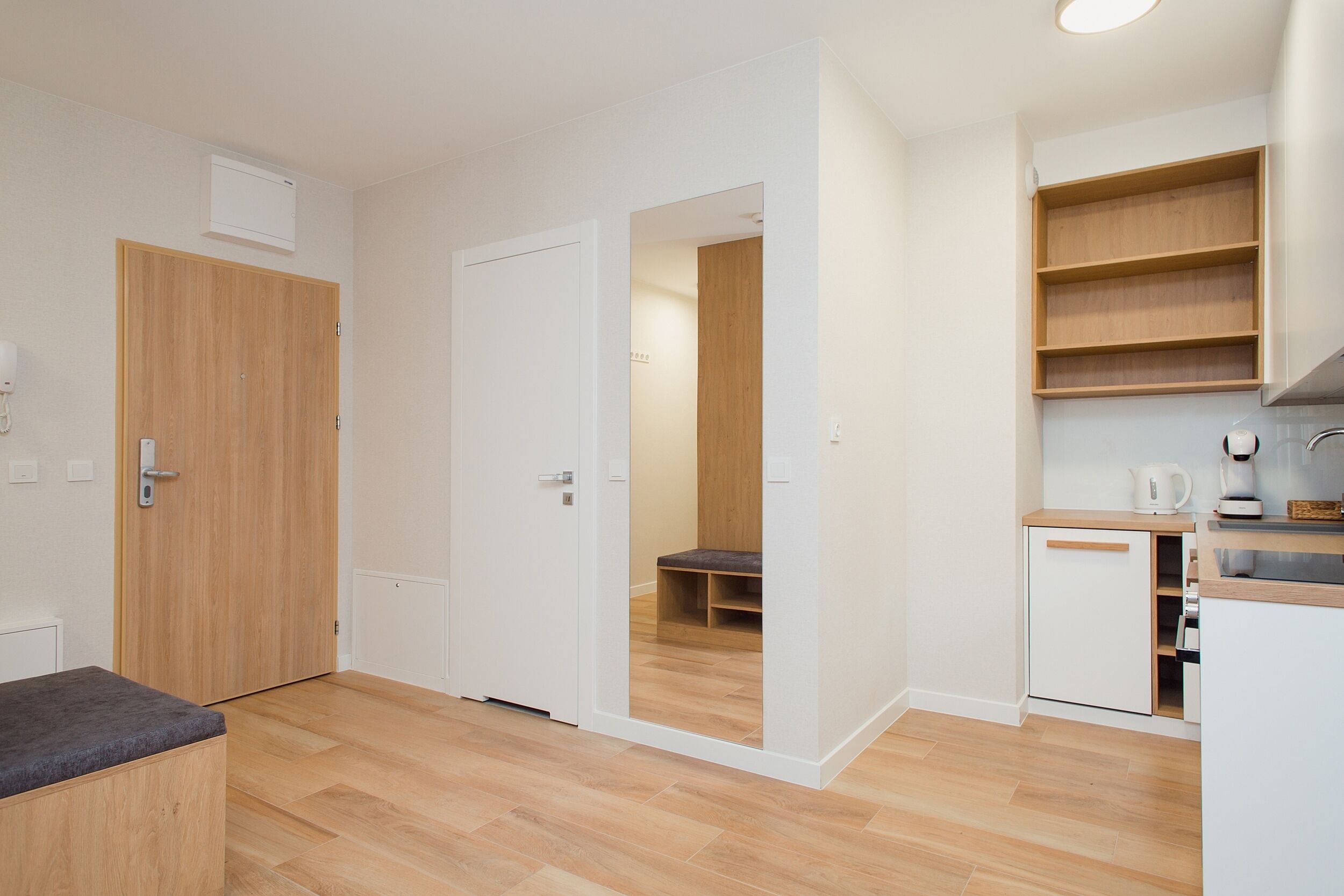 A modern apartment with a wooden floor, white walls, a kitchen with a coffee machine, and a bench in the living area.