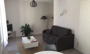 A living room with a grey sofa, a white coffee table with a plant, a floor lamp, and a TV on a stand.