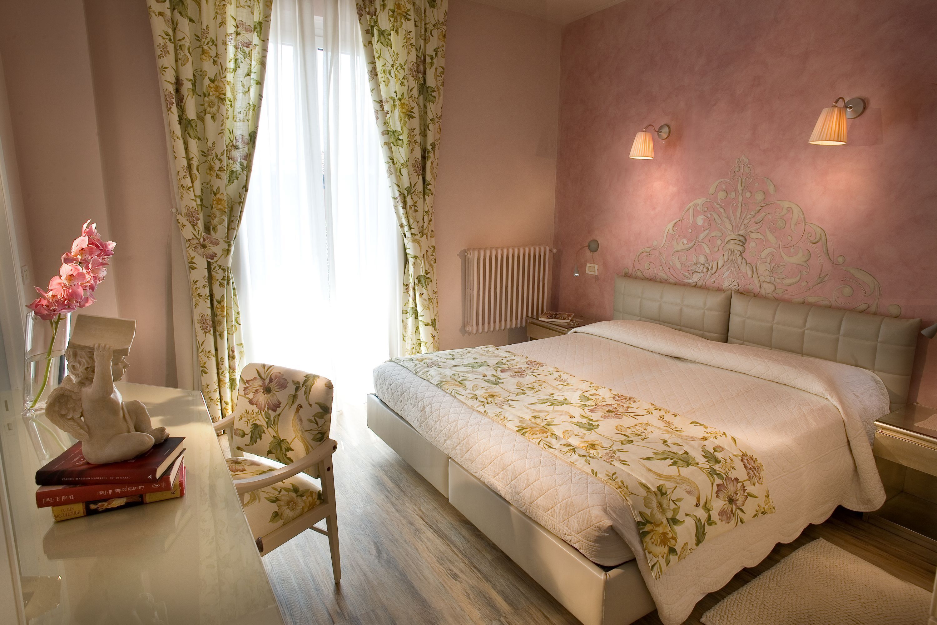Un dormitorio con una cama grande, una mesita de noche, una silla, un radiador y cortinas con estampado floral.
