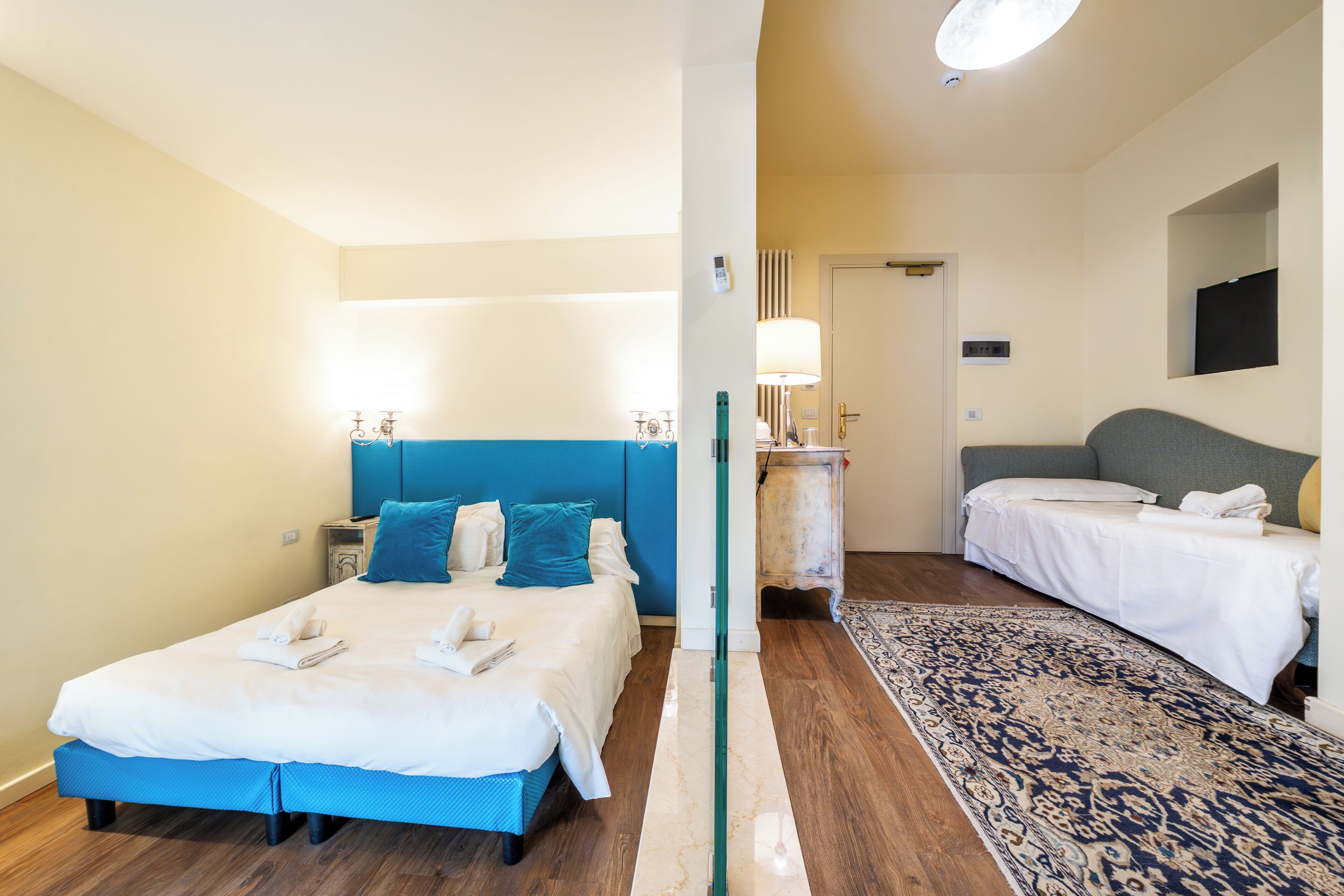 Une chambre d’hôtel avec un grand lit, un plus petit lit, une table de chevet, une lampe et un miroir au mur.