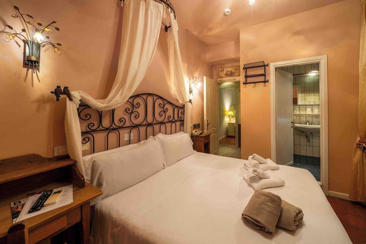 A bedroom with a four-poster bed, a bedside table with a lamp, and a bathroom visible through an open door.