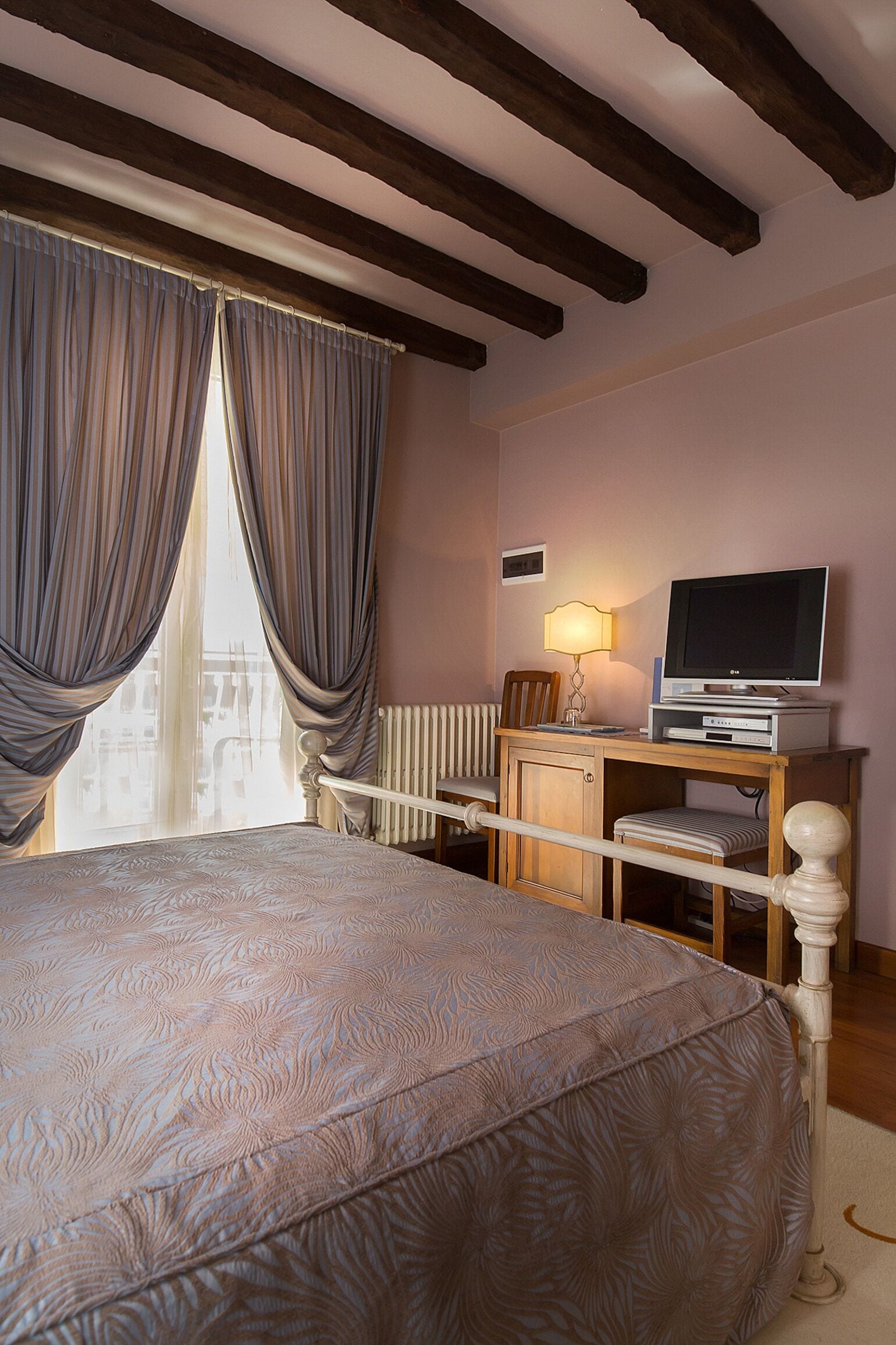 Une chambre à coucher avec un lit, un téléviseur posé sur un meuble en bois, une lampe et une fenêtre avec des rideaux.