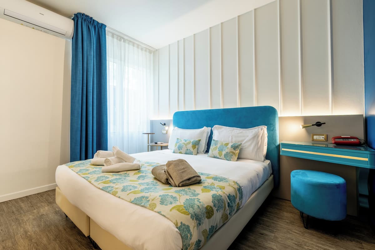 A hotel room with a bed, a desk, a chair, a nightstand, and a window with curtains.