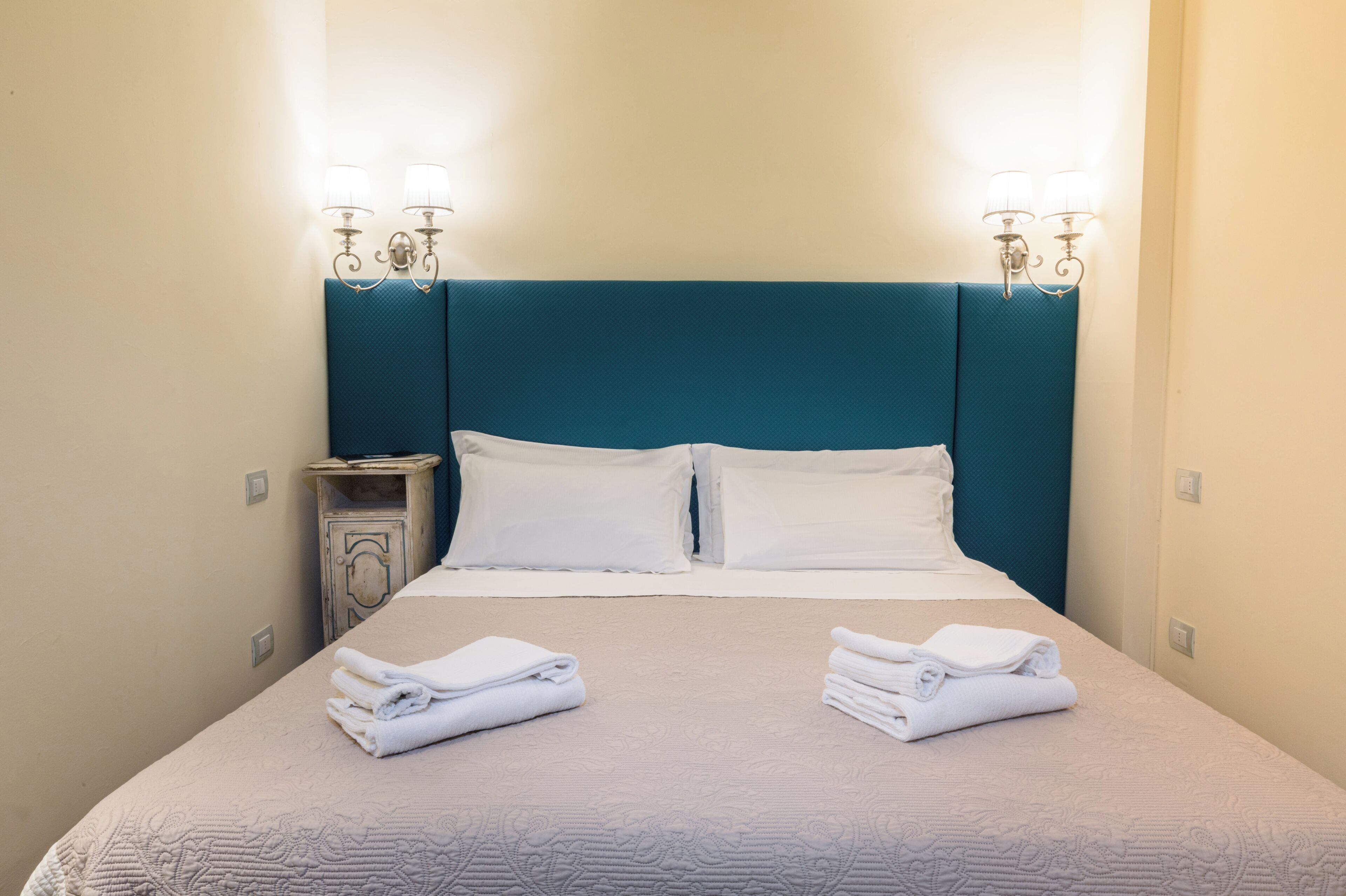 Una habitación de hotel con una cama, dos lámparas de pared, una mesita de noche y toallas dobladas.