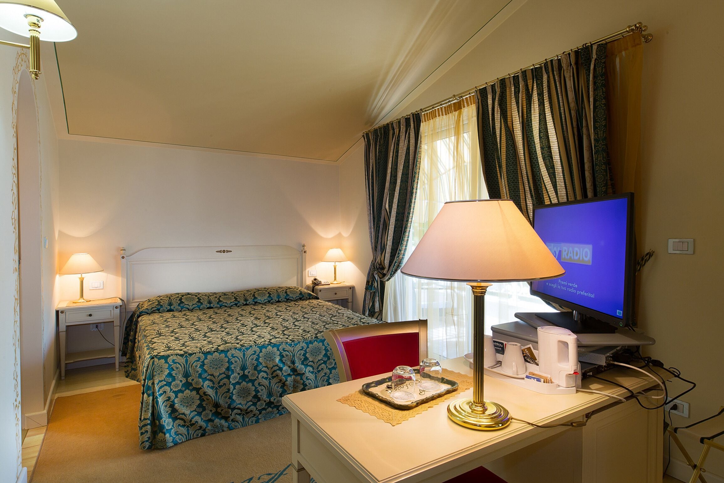 A hotel room with a bed, bedside lamps, a desk with a lamp, a television, and a window with curtains.