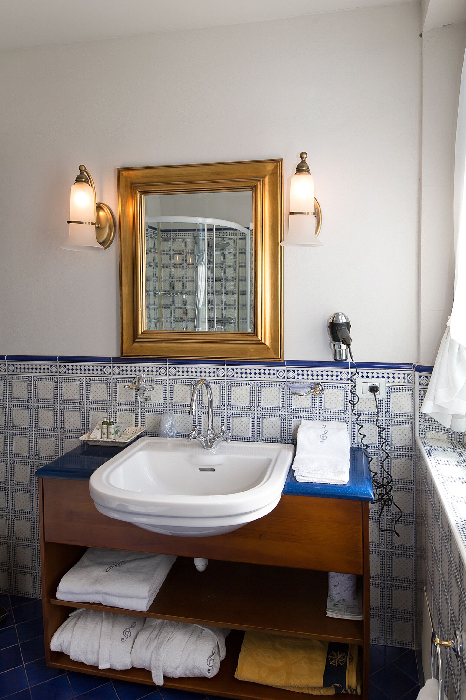 Un baño con un lavamanos blanco, un espejo con marco dorado y azulejos con diseño azulado.