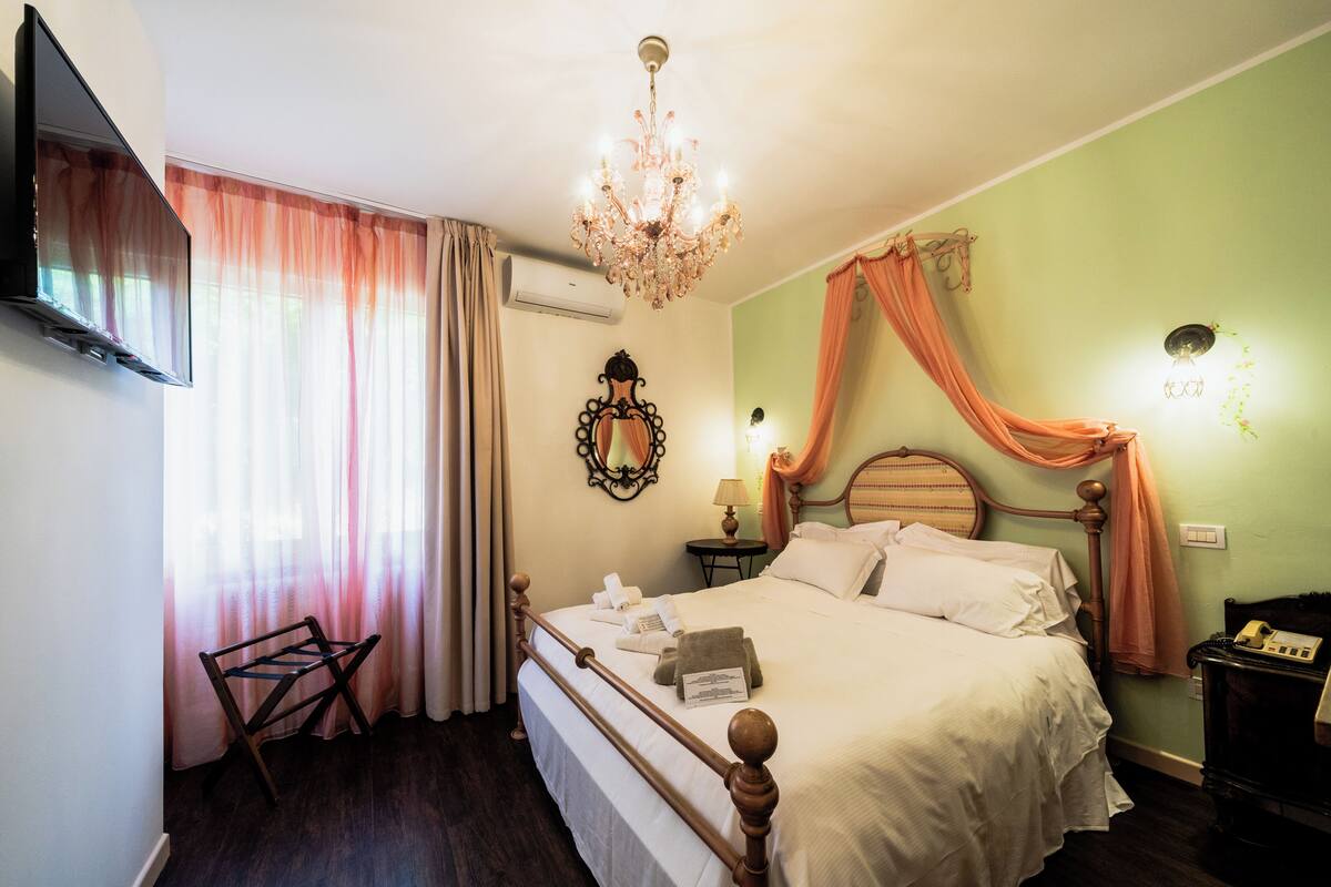A hotel room with a bed, a TV, a chandelier, a bedside table, a lamp, and a telephone.