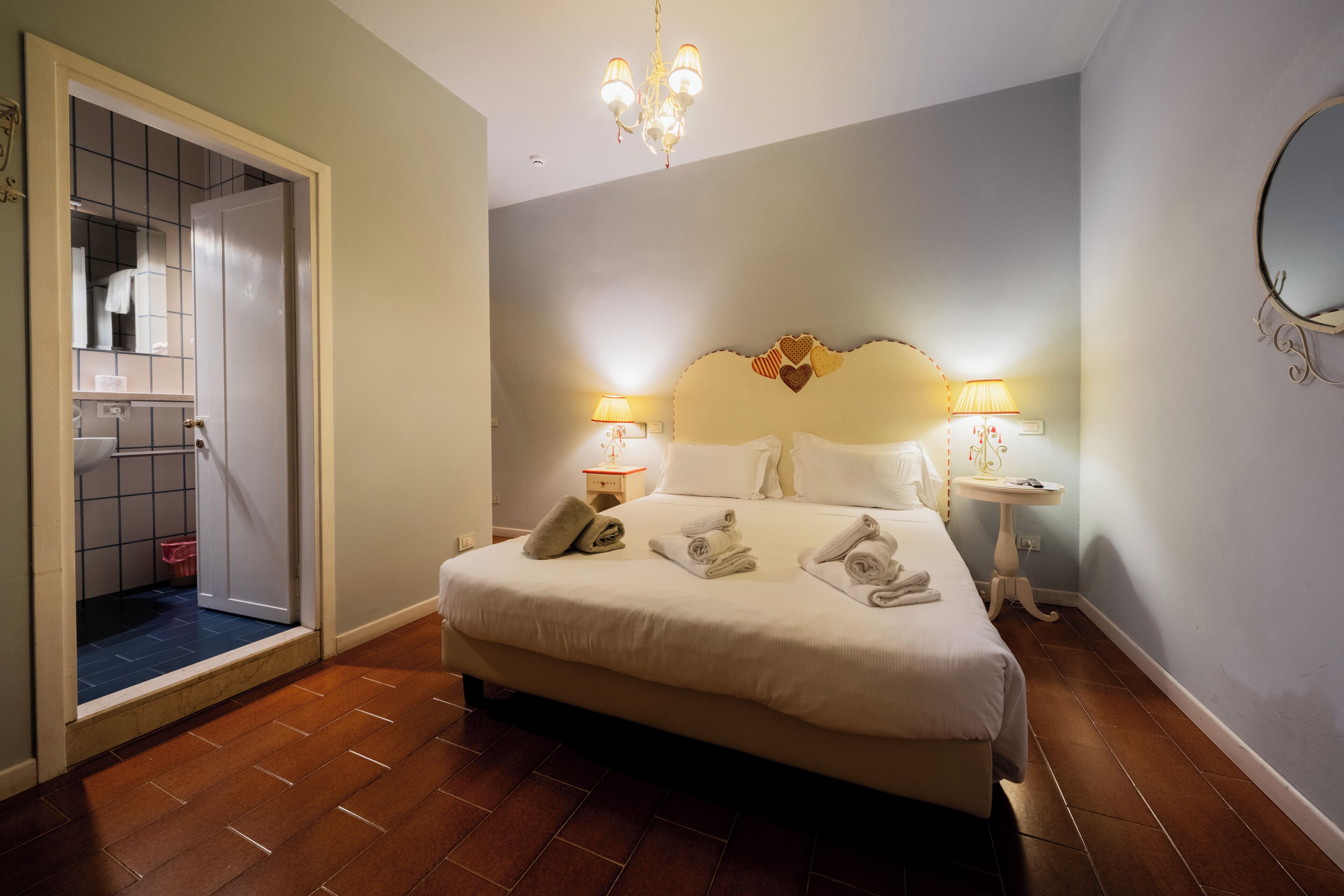 A hotel room with a bed, bedside lamps, a chandelier, and a bathroom visible through an open door.