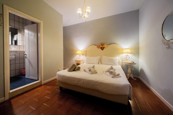 Habitación de hotel con cama, lámparas de noche, una araña y un baño visible a través de una puerta abierta.