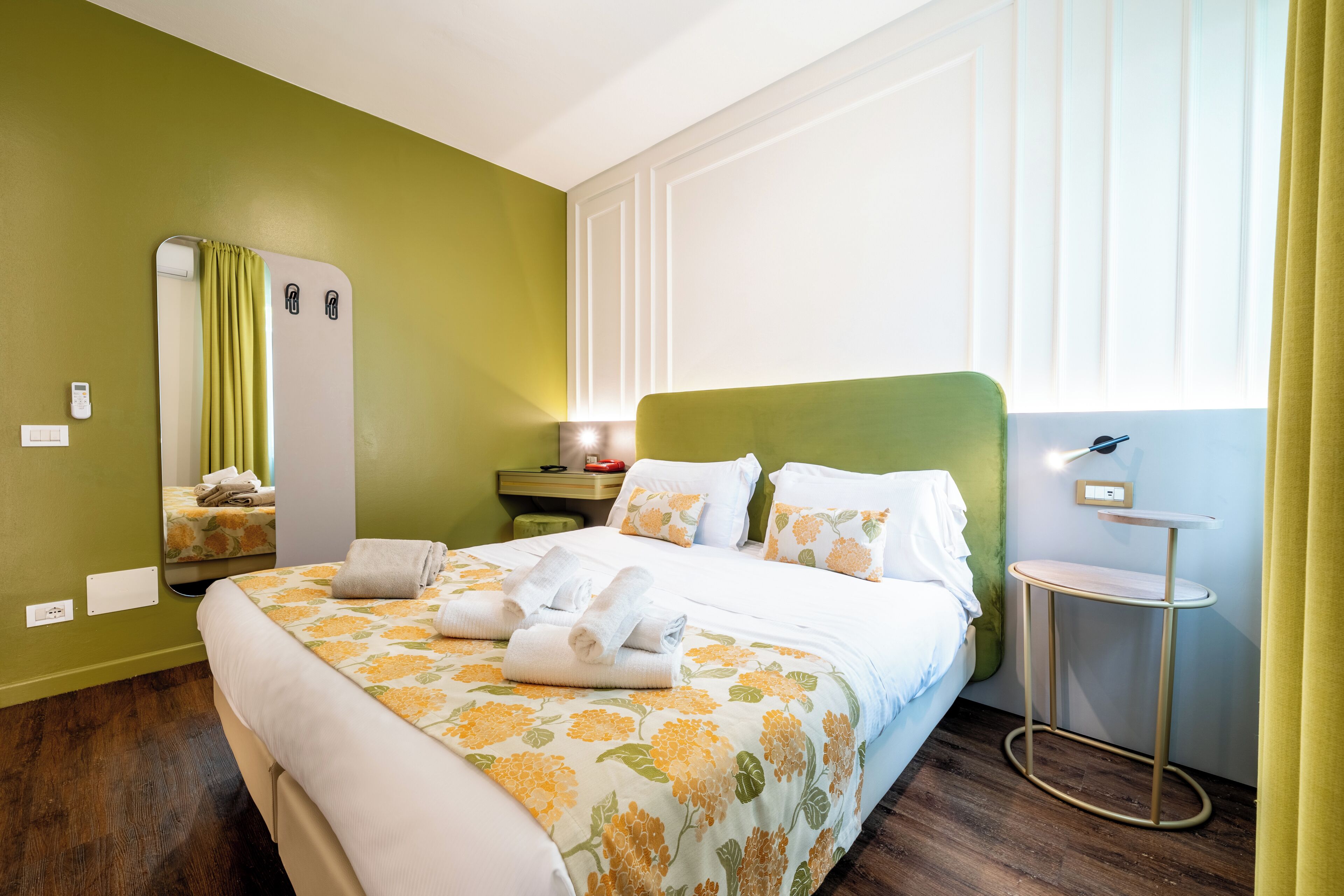A hotel room with a bed, a bedside table, a mirror, and a window with curtains.