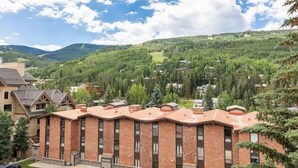 Un immeuble à plusieurs étages, doté d’une façade en briques et d’un toit plat, entouré d’une végétation luxuriante et de collines.