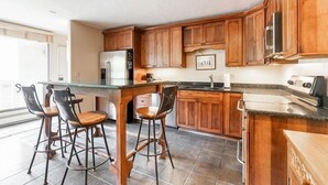 Une cuisine avec des armoires en bois, un réfrigérateur en acier inoxydable, un four à micro-ondes et une cuisinière à gaz. On y trouve trois tabourets de bar et un îlot central avec un dessus en verre.