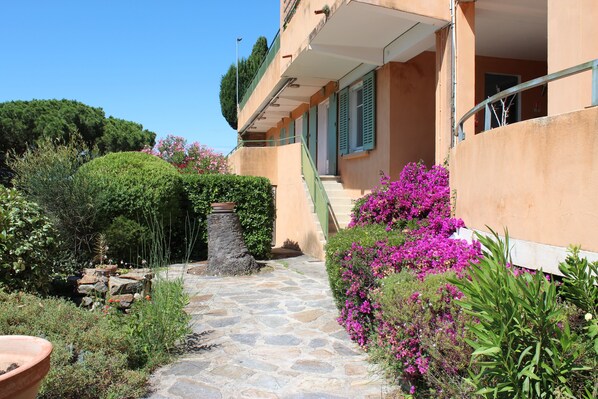 A stone pathway leading to a building with a balcony, surrounded by lush greenery and flowering plants.