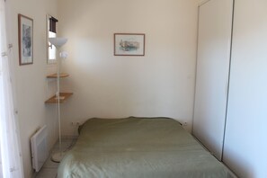 A small, sparsely furnished room with a bed, a radiator, a floor lamp, and two framed pictures on the wall.