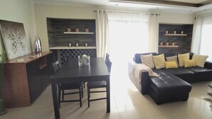 A modern living room with a black sofa, a dining table, and a kitchen area with shelves. - OURASOL 2 Bedroom Apartment in Santa Eulalia, Albufeira Centre  (Albufeira)