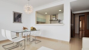 A modern dining area with a glass table, white chairs, and a chandelier. A kitchen is visible in the background through a large mirror. - Minimal, sea views apartment first line (Santa Margalida - Can Picafort/Son Serra Marina)