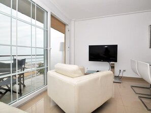 A modern living room with a large sliding glass door leading to a balcony, a flat-screen TV mounted on the wall, and a white sofa.