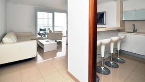 A modern living room with a sofa, a coffee table, and a dining area with chairs. There is a kitchen with a sink and bar stools. - Minimal, sea views apartment first line (Santa Margalida - Can Picafort/Son Serra Marina)