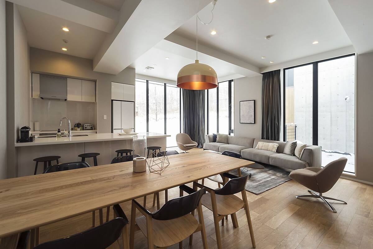 A modern living space with a wooden dining table, black chairs, a grey sofa, and a large window providing natural light.