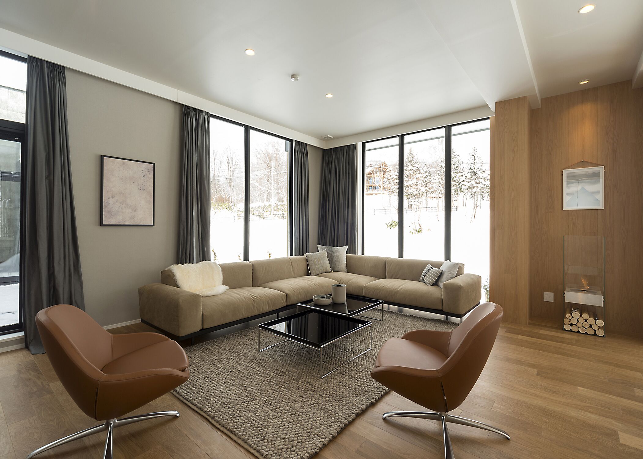 A modern living room with a beige sectional sofa, two brown chairs, a glass coffee table, and large windows with curtains.