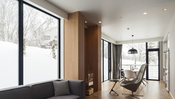 Una sala de estar moderna con un sofá gris, una zona de comedor con mesa y sillas, y ventanas amplias que ofrecen vistas a un paisaje nevado.