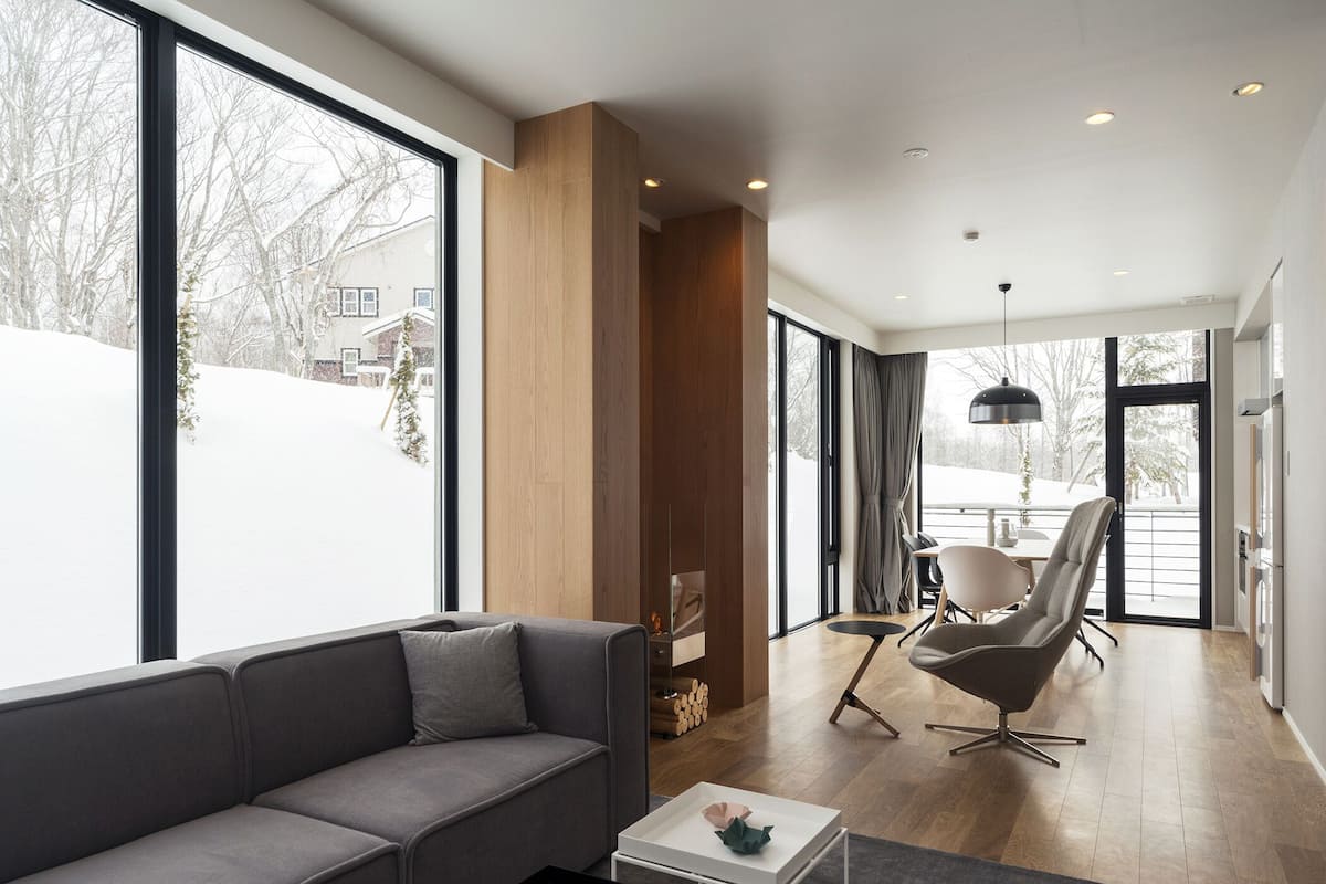 A modern living room with a grey sofa, a dining area with a table and chairs, and large windows offering a view of a snowy landscape.