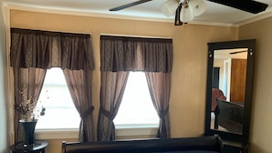 A bedroom with a bed, a ceiling fan, a window with curtains, a small table with a vase, and a mirror on the wall. - SHOTGUN HOUSE (Angleton)