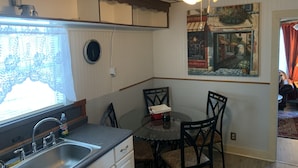 A kitchen with a sink, cabinets, and a dining table with chairs. There is a ceiling fan with lights, a window with lace curtains, and a wall-mounted clock. - SHOTGUN HOUSE (Angleton)