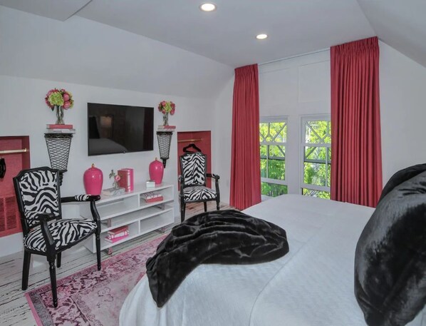 Un dormitorio con una cama grande, sillones estampados de cebra, un televisor y una ventana con cortinas rojas.
