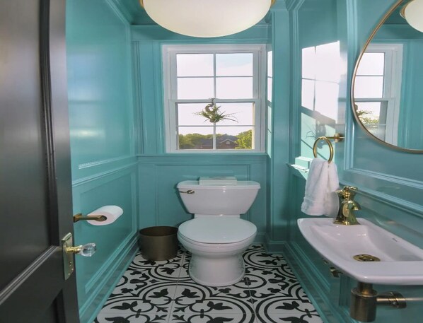 A bathroom with teal walls, a white toilet, a sink with a gold faucet, and a window with a plant.
