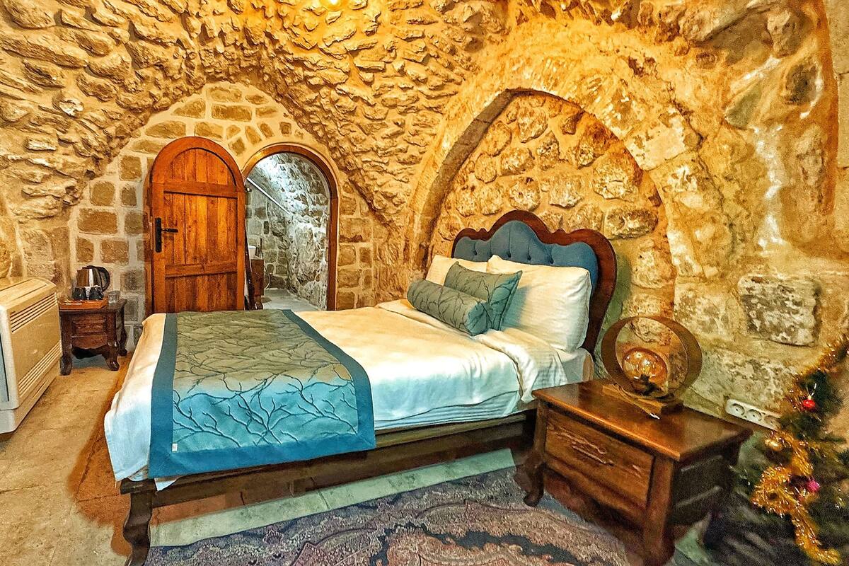 A bedroom with a stone wall, a wooden door, a bed with a blue bedspread, a nightstand, and a radiator.