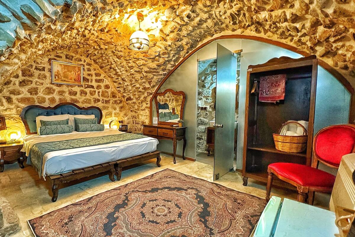 A bedroom with a stone ceiling, a bed with a wooden headboard, a red chair, and a mirror on the wall.