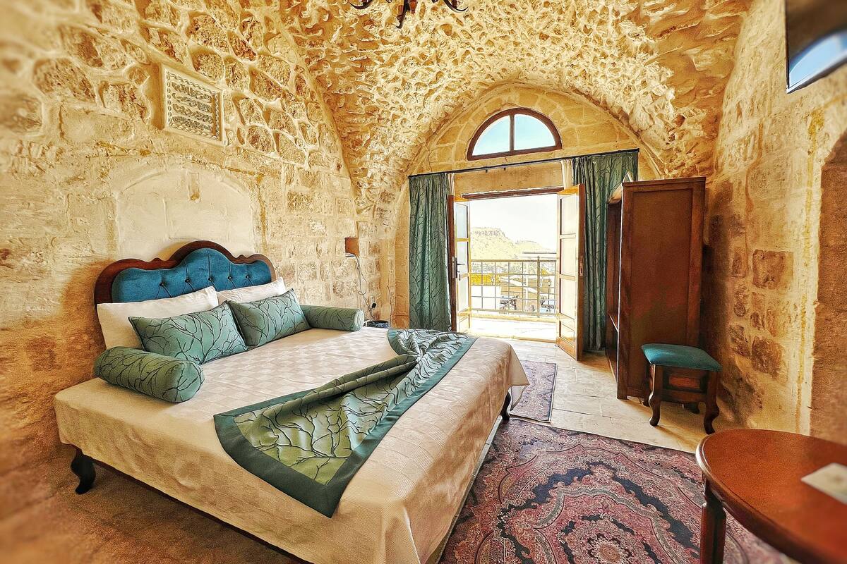 A bedroom with a stone wall, a bed with a blue headboard, a wooden chair, and a view through an open door.
