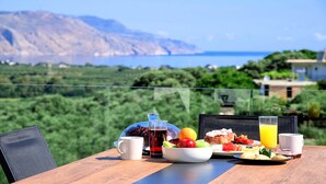 A table with breakfast items and a glass of orange juice, overlooking a scenic landscape with mountains and the sea.