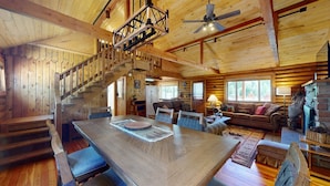 Un intérieur spacieux de chalet en bois, avec un escalier en bois, un coin repas, un salon doté d’une cheminée et une grande fenêtre laissant entrer la lumière naturelle.
