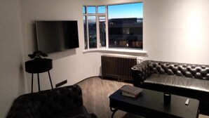 A modern living room with a leather sofa, a flat-screen TV, a small table, and a window with a city view.